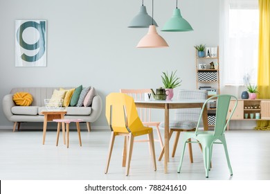 Modern Colorful Chairs At Dining Table Under Pastel Lamps In Living Room Interior With Pillows On Settee Against Wall With Poster