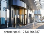 Modern commercial building entrance with a glass facade, revolving doors, and street reflections in a business district, sunny day with cars parked on the sidewalk