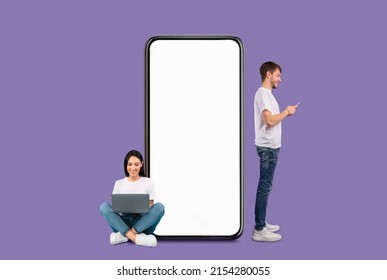 Modern Communication Concept. Cheerful Couple Chatting On Social Media, Smiling Guy Using Cell Standing Near Huge Big Smartphone With Blank White Screen, Lady Sitting On Floor With Pc, Purple Wall