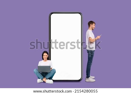 Modern Communication Concept. Cheerful Couple Chatting On Social Media, Smiling Guy Using Cell Standing Near Huge Big Smartphone With Blank White Screen, Lady Sitting On Floor With Pc, Purple Wall