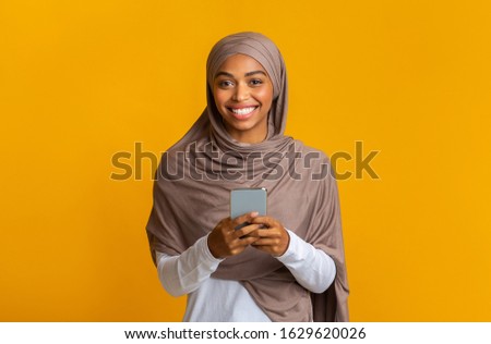 Modern Communication Concept. Portrait Of Cheerful Afro Muslim Girl In Hijab With Smartphone In Hands Looking At Camera, Yellow Background, Copy Space
