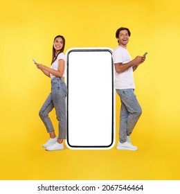 Modern Communication. Full Body Length Of Happy Cheerful Couple Leaning On Big Huge Smartphone With White Empty Blank Screen Using 2 Cell Phones, Chatting On Social Media Standing On Orange Background