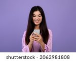 Modern communication. Happy armenian woman browsing on smartphone, messaging or chatting with friends, scrolling social networks feed over purple background, studio shot