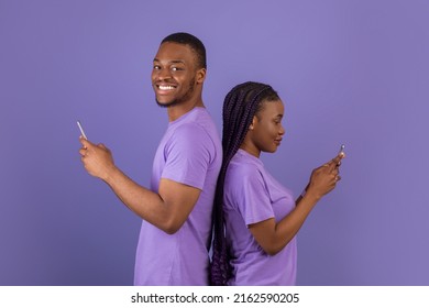 Modern Communication. Profile Side View Portrait Of Cheerful Positive Black Couple Standing Back To Back, Holding And Using Their Mobile Phones And Chatting Online, Guy Looking At Camera, Purple Wall