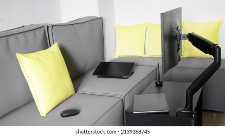 Modern computer monitor is mounted on a small table with a gas-lift metal swivel arm. Next to a laptop, an electronic tablet, a computer mouse and a large loft sofa with yellow pillows. Daylight
