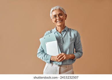 Modern confident beautiful gray-haired mature business woman in blue blouse and glasses holding laptop and work folders while standing on beige background and smiling. - Powered by Shutterstock