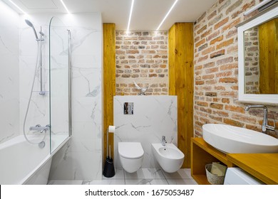Modern contemporary loft interior of bathroom. White bath and sink. Brick wall. Toilet and bidet.