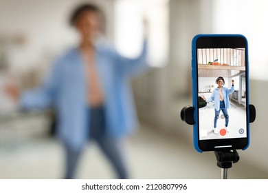 Modern Content Creator. Positive African American young lady in headset filming video blog for social network, using cell phone on tripod to broadcast in living room, selective focus on device screen
