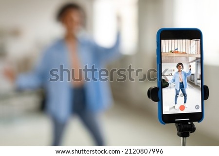 Modern Content Creator. Positive African American young lady in headset filming video blog for social network, using cell phone on tripod to broadcast in living room, selective focus on device screen