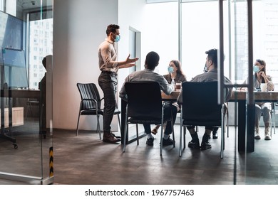 Modern Corporate Meeting Concept. Young Businessman In Medical Face Mask Giving Speech During Meeting With Coworkers In Office, Standing At Table In Board Room, Explaining Strategy And Management