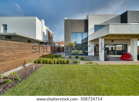 Modern country house with the large lawn and a wooden fence. In front of the house there is a covered terrace with a lounge zone. On the lawn there is a trees and flowerbeds. It is sunny. Outside.