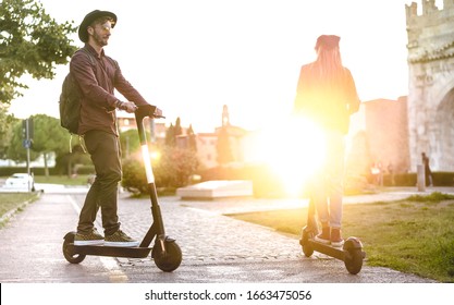 Modern Couple Using Electric Scooter In City Park - Milenial Students Riding New Ecological Mean Of Transport - Green Eco Energy Concept With Zero Emission - Bright Warm Filter With Sunshine Halo