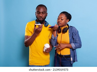 Modern Couple Using Speaker On Smartphone To Record Message, Wearing Headset Over Blue Background. Boyfriend And Girlfriend Holding Mobile Phone To Use Microphone, Making Speech Online.