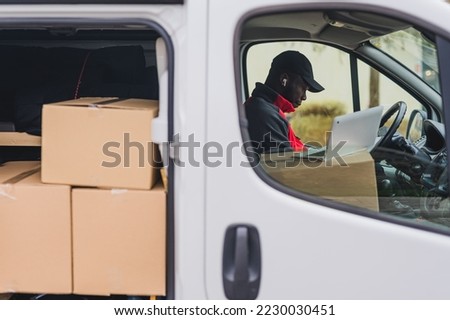 Modern courrier. Online shopping package delivery concept. White delivery van with opened side doors revealing many undelivered packages. Adult Black deliveryman in dark-colored hat and red jacket