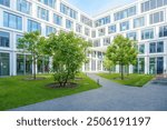 modern courtyard enclosed by a white building with large glass windows. The courtyard features a well-maintained green lawn with several young trees planted in circular soil beds