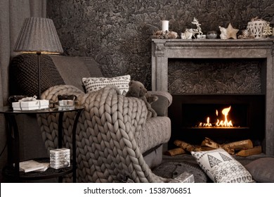 Modern Cozy Interior In Gray Shades With An Armchair In Front Of Fireplace. On The Chair There Are Nice Knitted Blanket, Cushions And Teddy Bear. A Lamp And Gifts On The Table. 