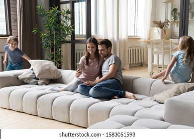 In modern cozy living room full family young couple sit on couch hold cellphone make call, purchasing using e-commerce while kids running play tag game, homeowners weekend activity at new home concept - Powered by Shutterstock