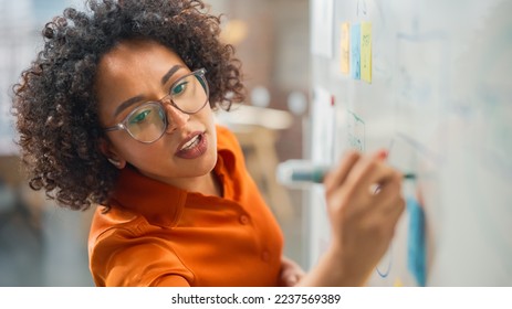 Modern Creative Agency Meeting: Confident Black Female Engineer Uses Whiteboard, Makes Report to a Group of Engineers, Managers Talks, and Shows Statistics, Growth and Analysis Information - Powered by Shutterstock