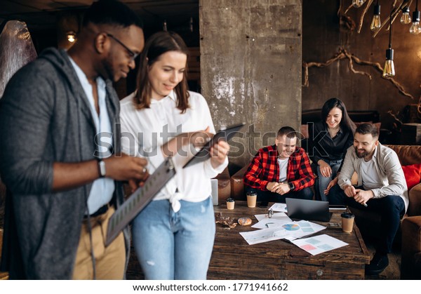 Modern creative
office.  Successful colleagues are divided into groups to develop
new ideas. African-American guy and a Caucasian girl look at a
tablet and discuss new
startup