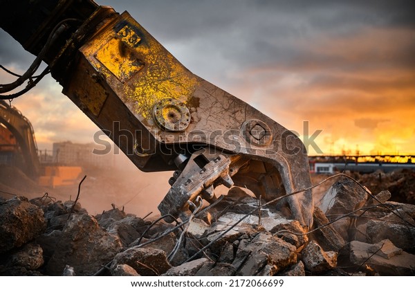 Modern crusher on excavator rigging dismantles\
reinforced concrete beam for recycling at demolition site at sunset\
light closeup