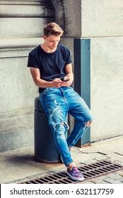 Modern Daily Life. Young American Man With Little Beard Texting On Cell Phone In New York, Wearing Black T Shirt, Destroyed Jeans, Fashionable Shoes, Sitting On Pillar In Corner Of Vintage Street.