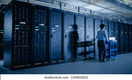In the Modern Data Center: IT Engineer Doing Maintenance and Diagnostics. Pushes Cart alongside Server Racks. Planned System Equipment Update and Maintanence.