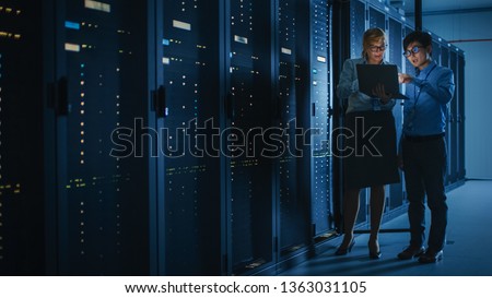 In the Modern Data Center: Engineer and IT Specialist Work with Server Racks, Talking, Doing System Maintenance Check with Laptop Computer. Concept of Diagnostics of the Database, Cybersecurity Check