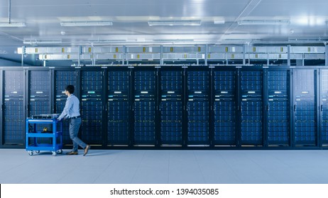 In the Modern Data Center: IT Technician Working with Server Racks, Pushes Cart Between Rows of Server Racks. On a Pushcart New Hardware for System Update. Engineer Doing Maintenance and Diagnostics.