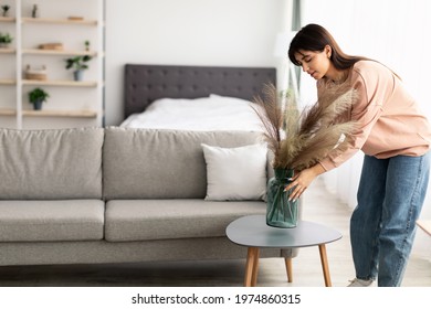 Modern Decoration Concept. Casual young lady putting glass vase with dried flowers on tea table. Millennial woman decorating her modern apartment and bedroom or living room with pampas grass