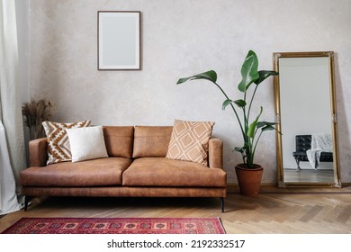 Modern design of living room with brown eco leather couch, soft cushions, mirror with golden frame, copy space picture frame on wall and houseplant in pot
