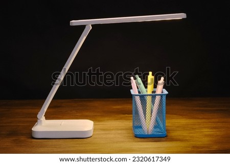 Modern design rechargeable desk lamp illuminates a ballpoint pen holder against a dark background. The concept of study or office work.