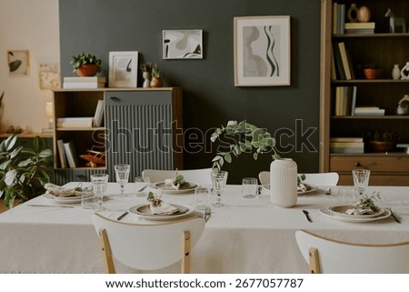 Modern dining room table set for meal with glassware, plates, cloth napkins, and ceramic vase with greenery, contemporary wall art and plants decorating background shelves