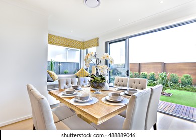 Modern Dining Table Set Up Indoors With Outdoor Garden View In A Luxury House, Shiny Wooden Table In Brown Color Has Ceramic Cups And Dishes With A Glass Vase And Flowering Plant Decoration, 