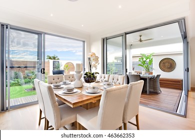 Modern Dining Table Set Up With Outdoor Patio Decoration At Daylight Including Close Up Of A Wooden Table With Chairs, There Are Ceramic Mugs With A Flowering Vase On It, There Are Glass Doors 