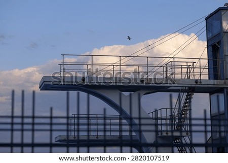 Modern diving platform complex extending into the open sky, featuring multiple levels and metal railings against a backdrop of clouds, with a bird flying freely above the structure