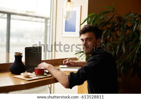 Modern dressed man sit in cafe with notebook and look to the camera with hands on laptop. Freelancer wait for the client for new job and use free wi-fi to surfing internet pages