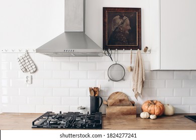 Modern eclectic kitchen interior with pumpkins. White brick wall with metro tiles, peg rails and oil painting. Wooden countertop, hood and gas stove. Scandinavian home design. Thanksgiving, Halloween.
