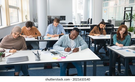 Modern Education Concept. Portrait Of Diverse Group Of Students Sitting At Desks In Classroom At University, Taking Test, Entrance Examination Or Writing Notes In Notebook, Selective Focus