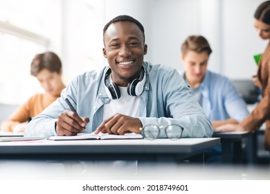 Modern Education Concept. Portrait Of Smiling African American Male Student Sitting At Desk In Classroom At University, Taking Test Or Writing Notes In His Notebook, Looking Posing At Camera