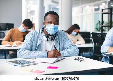 Modern Education During Pandemic Concept. Portrait of african american male student sitting at desk in classroom at university, wearing protective medical mask, writing in notebook, looking at camera