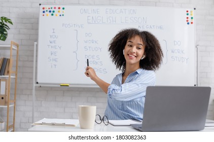 Modern education remotely. Cheerful young african american woman points to blackboard and explains rules of english online