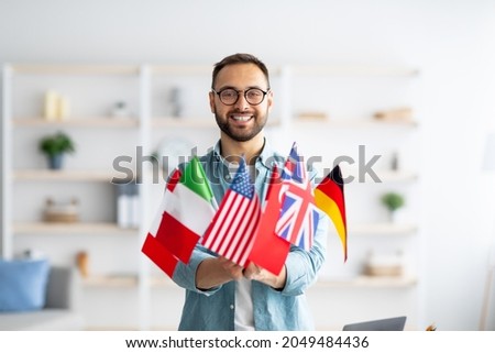 Modern education and student exchange. Cheerful young Caucasian guy holding bunch of different flags at home. Happy millennial man advertising foreign languages school, emigrating overseas
