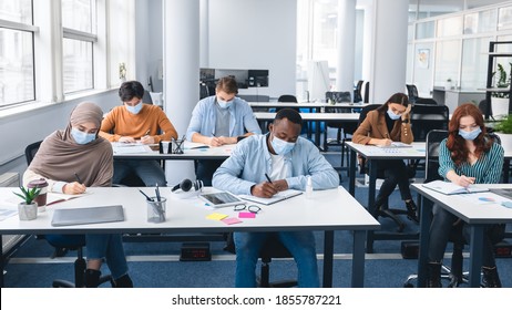 Modern Education And Studying During Pandemic Concept. Diverse Group Of International Students Sitting At Desks In Classroom At High School, Wearing Protective Medical Mask, Writing In Notebooks