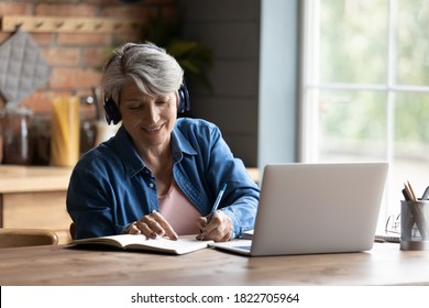 Modern elderly grey-haired 60s woman in headphones take online course or training on computer at home. Smart mature Caucasian female in earphones watch webinar make note study distant on laptop.