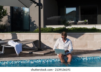 A modern elderly man enjoys the pool while working on his laptop next to a modern luxury house. Selective focus 