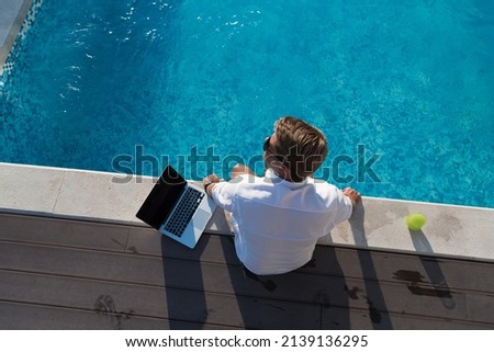 A modern elderly man enjoys the pool while working on his laptop next to a modern luxury house. Selective focus 