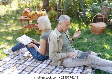 Modern Elderly Spouses Resting In Garden, Man Using Laptop Computer While His Wife Reading Book, Sitting Back To Back On Blanket. Retired Couple Spending Time Outdoors At Spring Day