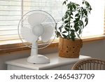 Modern electric fan with houseplant on commode at home