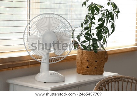 Modern electric fan with houseplant on commode at home