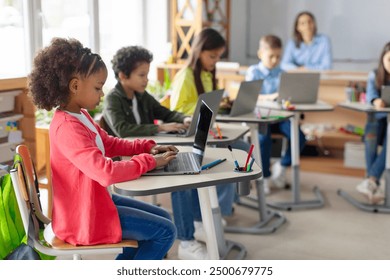 Modern elementary education concept. Group of diverse school children sitting at desks with laptops, studying, using digital gadgets - Powered by Shutterstock